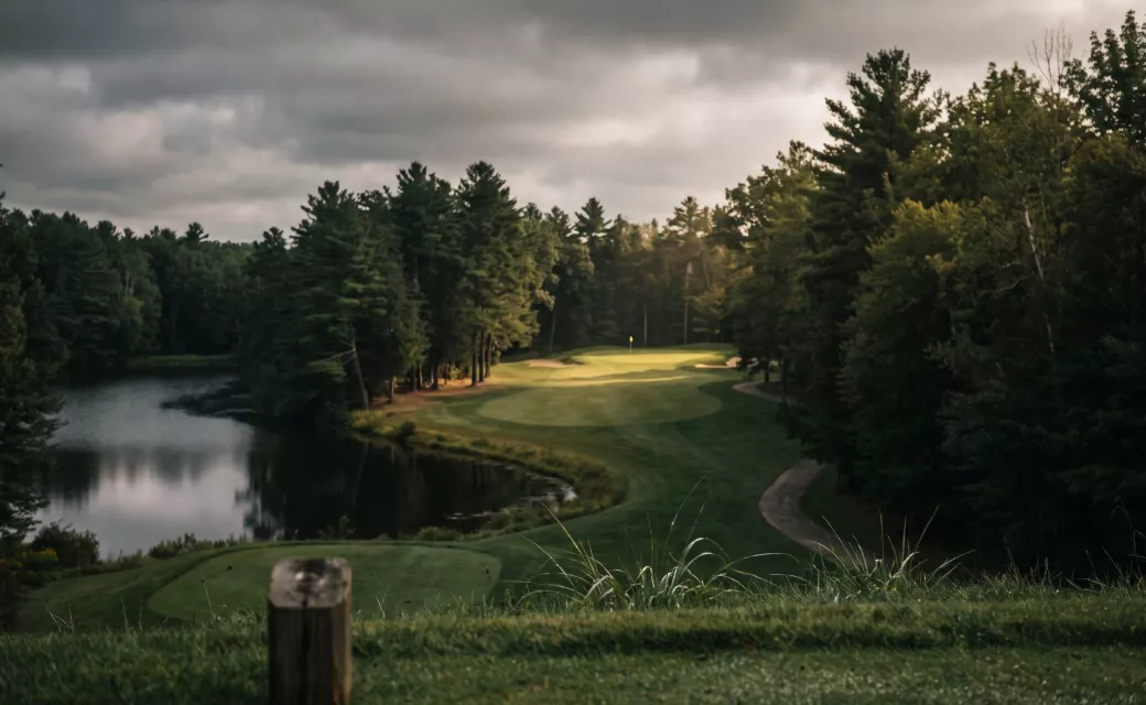 Trou exigeant vu du départ : fairway étroit entre les arbres et plan d’eau sous un ciel chargé